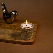 Decorative glass candle burning on wooden tray with gold bird figurine, brown background