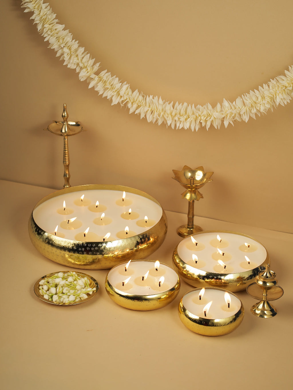 Brass urli with floating candles, jasmine flowers, and traditional brass lamps on beige background