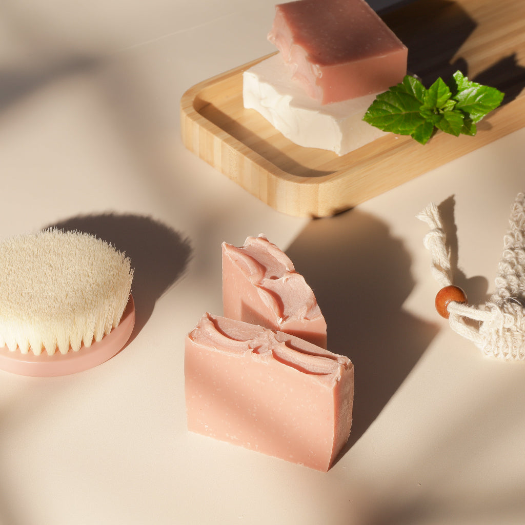Handmade pink and white soap bars on bamboo tray with body brush, mint leaf, and rope scrubber