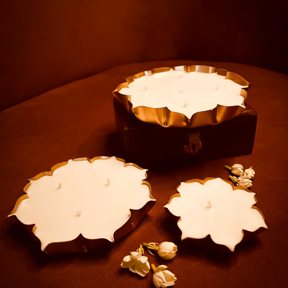 Three white floral-shaped candles in gold holders with jasmine flowers on a brown surface