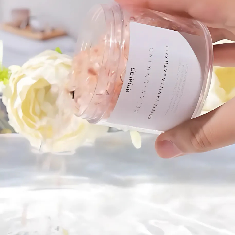 Hand pouring amarae coffee vanilla bath salt into water, white flower in background.
