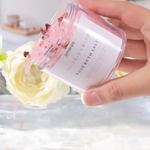 Load image into Gallery viewer, Hand holding Amaraa rose bath salt jar over water, white rose flower in background