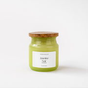 A green Jasmine Noir scented candle in a glass jar with a wooden lid on a white background.