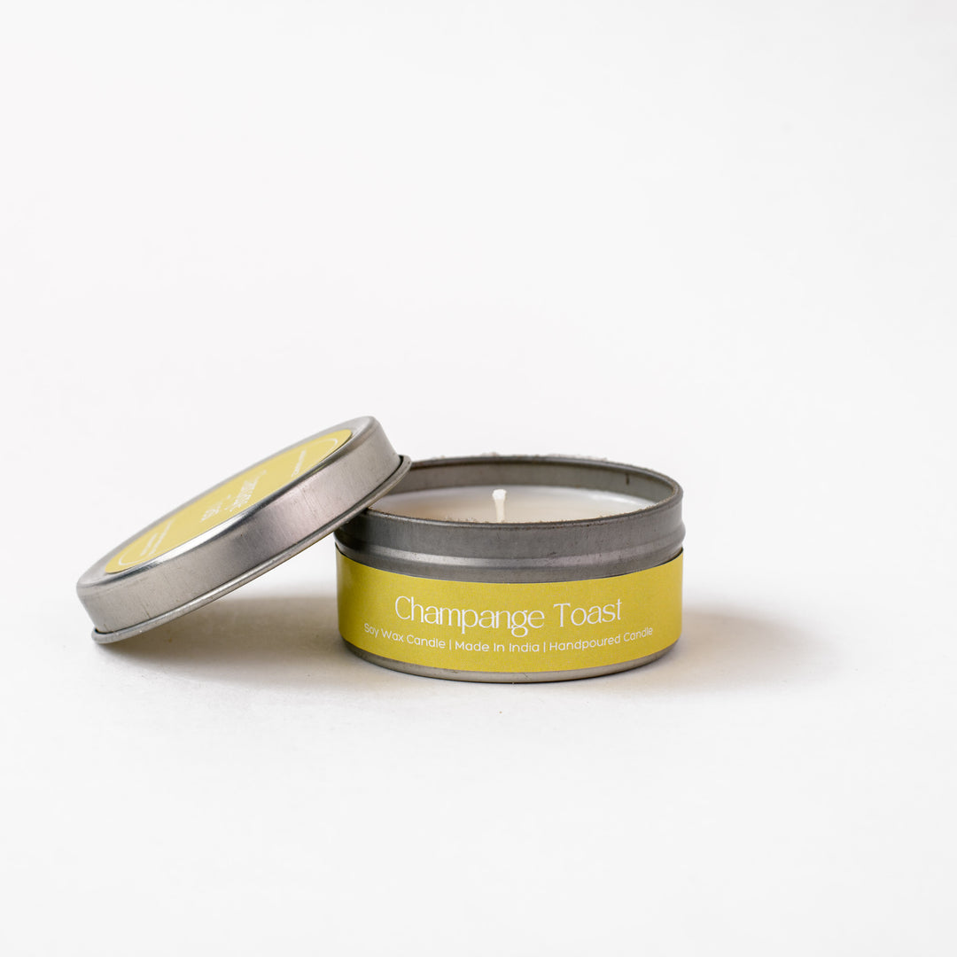 Champagne Toast soy wax candle in silver tin with yellow label, lid resting aside on white background