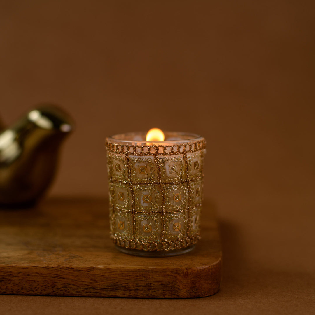 Decorative gold glass candle holder with lit candle on wooden board, brown background