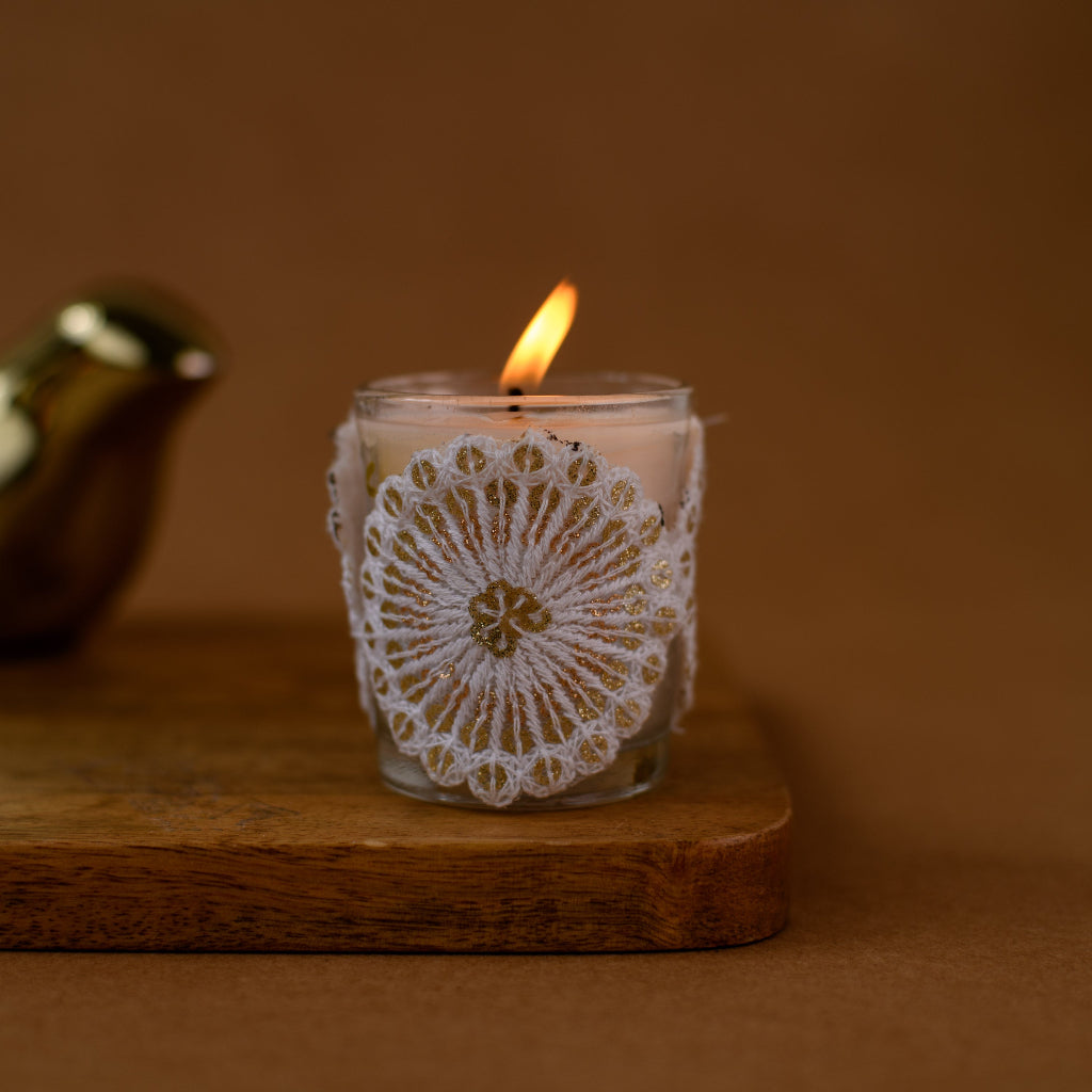 Handmade white lace-wrapped glass candle burning on wooden board, brown background