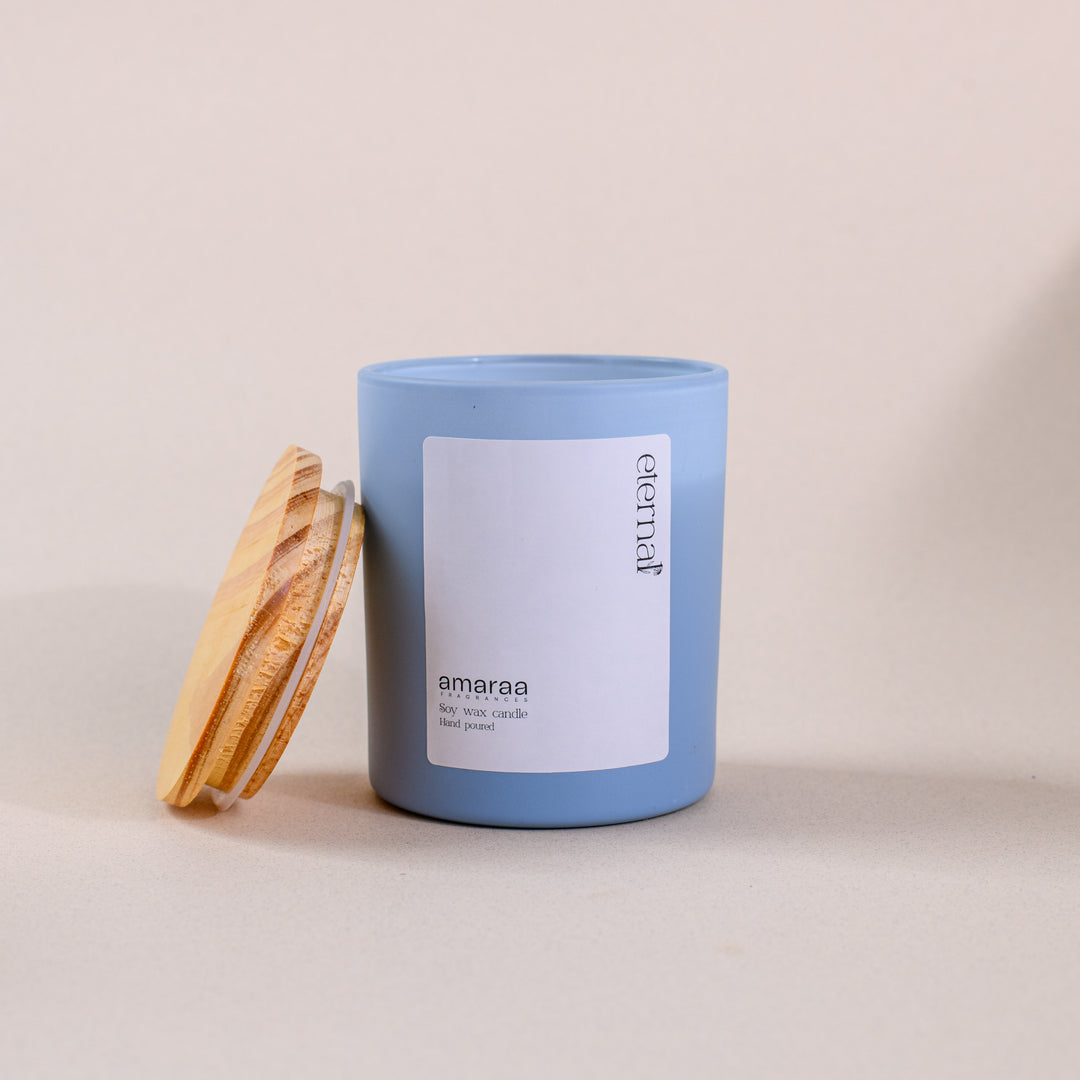 Blue soy wax candle in glass jar with wooden lid, labeled 'eternal amaraa', on neutral background