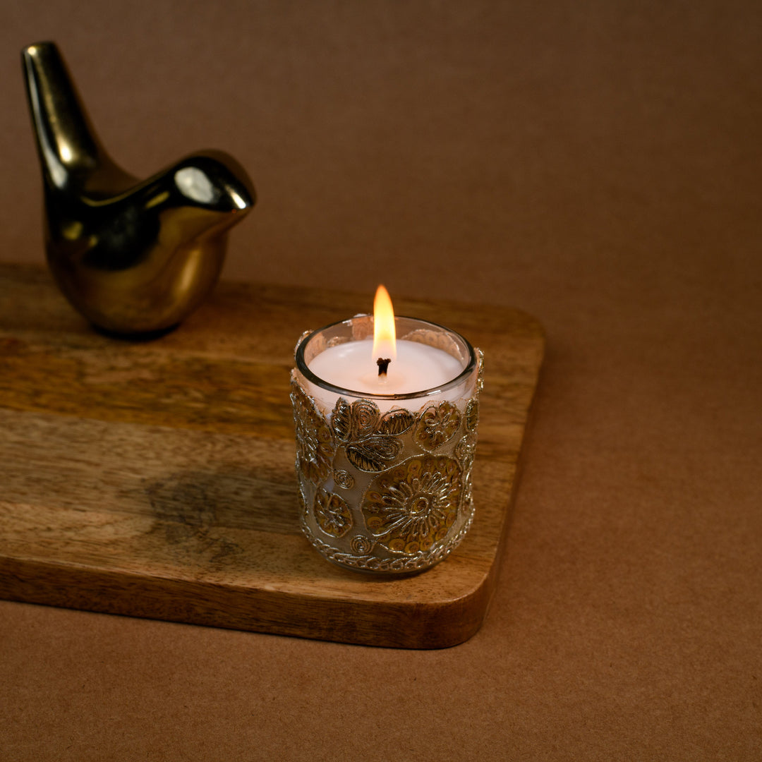 Decorative glass candle burning on wooden tray with gold bird figurine, brown background
